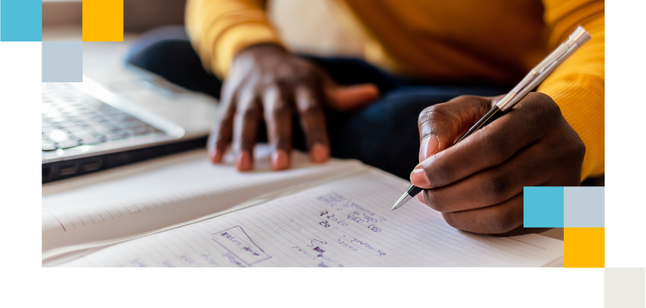 Close-up of a learner writing notes beside a laptop, preparing for a task.
