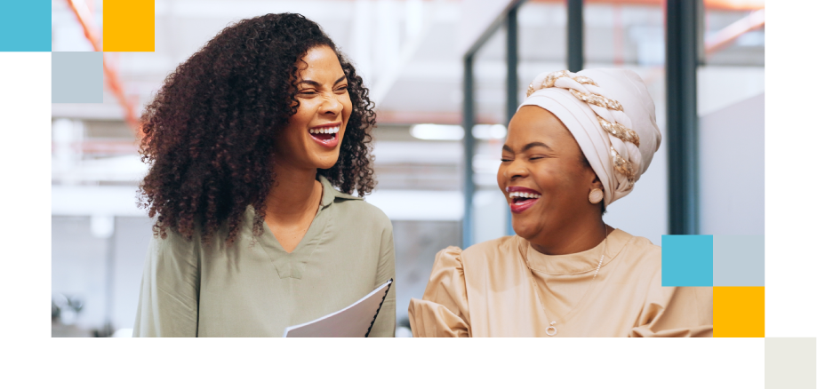 Two colleagues laugh together in an office corridor; one carries a notebook, the other wears a light headscarf.